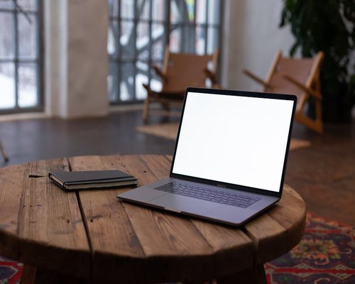 A closed laptop on a wooden table next to a notebook in a bright room
